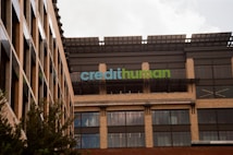 A modern building facade with large windows and a sign displaying 'credithuman'. The structure has a combination of beige brick, glass, and metal elements. Trees are partially visible in the foreground.