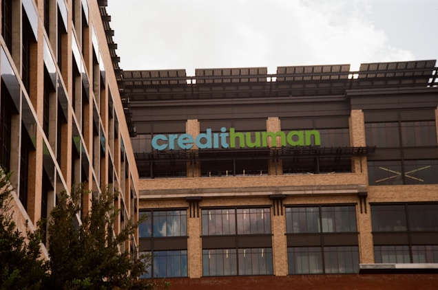 A modern building facade with large windows and a sign displaying 'credithuman'. The structure has a combination of beige brick, glass, and metal elements. Trees are partially visible in the foreground.