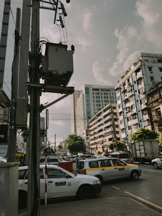 An urban street scene featuring a busy intersection filled with cars and taxis. Tall buildings line the street, contrasting with the cloudy sky above. Power lines and a utility pole are in the foreground, emphasizing the city's infrastructure.
