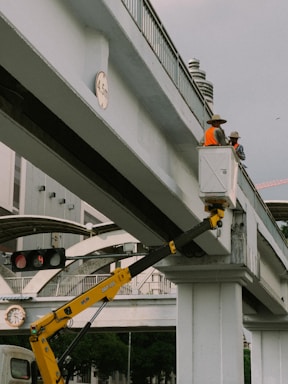 Two construction workers in bright orange safety vests and hats are standing on a raised platform attached to a crane arm, positioned on an elevated concrete bridge. The surroundings include urban infrastructure such as traffic lights and a building in the background. A height restriction sign indicating 4.5 meters is visible on the bridge.