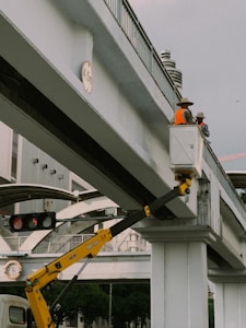 Two construction workers in bright orange safety vests and hats are standing on a raised platform attached to a crane arm, positioned on an elevated concrete bridge. The surroundings include urban infrastructure such as traffic lights and a building in the background. A height restriction sign indicating 4.5 meters is visible on the bridge.