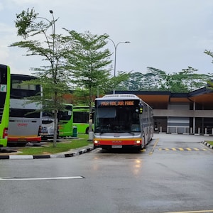 A modern bus station features multiple green and white buses parked under overcast skies. The central bus displays a message, 'YOUR RIDE, OUR PRIDE' on its front display panel. The area is surrounded by neatly maintained small trees, and the pavement is wet, suggesting recent rain.