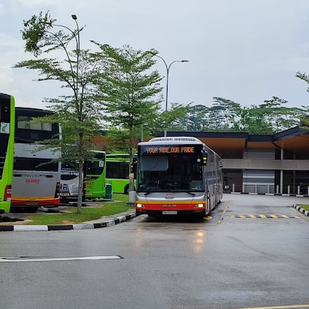 A modern bus station features multiple green and white buses parked under overcast skies. The central bus displays a message, 'YOUR RIDE, OUR PRIDE' on its front display panel. The area is surrounded by neatly maintained small trees, and the pavement is wet, suggesting recent rain.