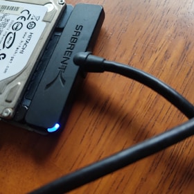 An internal hard drive connected to an external SATA to USB adapter, with a blue LED light indicating power, placed on a wooden surface. A black cable extends from the adapter.