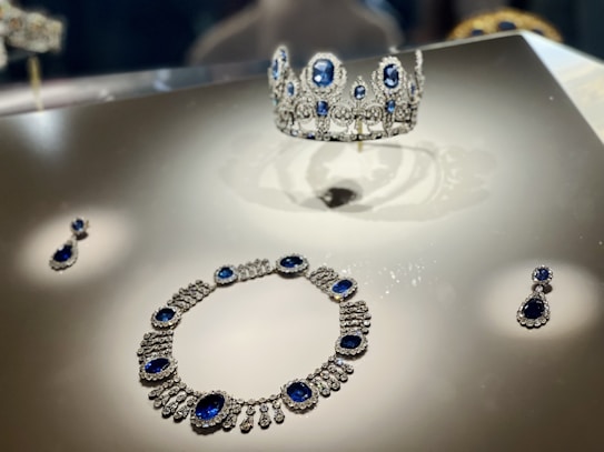 A display featuring a set of luxurious jewelry composed of a tiara, necklace, and earrings. Each piece is adorned with large, deep blue gemstones surrounded by intricate diamond settings, reflecting under the display lights.