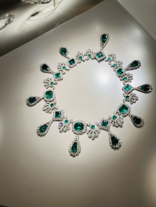 An elegant necklace encrusted with numerous emerald gemstones and surrounded by sparkling diamonds, arranged in an intricate pattern. The design features various shapes including teardrops and circles all beautifully set in a lustrous metal base.