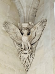 An intricately carved stone sculpture depicting a majestic eagle with outstretched wings. The eagle is adorned with detailed feathers and perched amidst ornate leaves and decorative elements, integrated into a classical architectural setting.