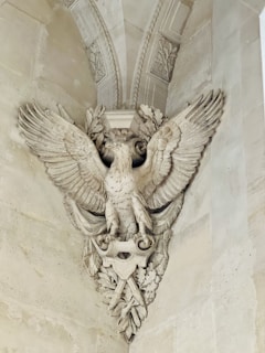 An intricately carved stone sculpture depicting a majestic eagle with outstretched wings. The eagle is adorned with detailed feathers and perched amidst ornate leaves and decorative elements, integrated into a classical architectural setting.