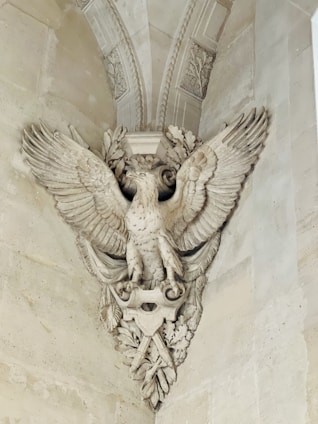 An intricately carved stone sculpture depicting a majestic eagle with outstretched wings. The eagle is adorned with detailed feathers and perched amidst ornate leaves and decorative elements, integrated into a classical architectural setting.
