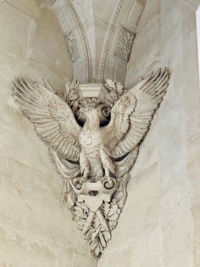 An intricately carved stone sculpture depicting a majestic eagle with outstretched wings. The eagle is adorned with detailed feathers and perched amidst ornate leaves and decorative elements, integrated into a classical architectural setting.