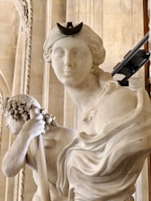 A close-up of two classical marble sculptures, featuring detailed facial expressions and intricate carving. One sculpture depicts a figure with a crescent-shaped headpiece, while the other holds a staff or rod. The background consists of ornate architectural details, suggesting a museum setting.