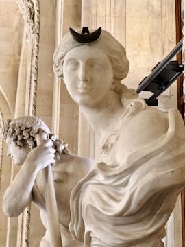A close-up of two classical marble sculptures, featuring detailed facial expressions and intricate carving. One sculpture depicts a figure with a crescent-shaped headpiece, while the other holds a staff or rod. The background consists of ornate architectural details, suggesting a museum setting.