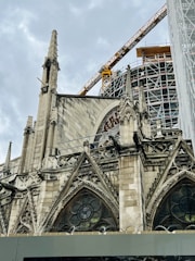 An ornate Gothic-style cathedral with pointed arches and stained glass windows is under renovation. A large construction crane and scaffolding are visible, indicating ongoing restoration work. The sky is overcast, enhancing the dramatic appearance of the architectural details.