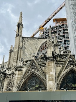 An ornate Gothic-style cathedral with pointed arches and stained glass windows is under renovation. A large construction crane and scaffolding are visible, indicating ongoing restoration work. The sky is overcast, enhancing the dramatic appearance of the architectural details.