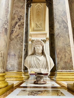 A marble bust of a bearded man draped in classical attire is elegantly positioned between ornate marble columns. The architecture surrounding the sculpture is richly decorated with intricate carvings and gold accents that reflect the grandeur of the setting.