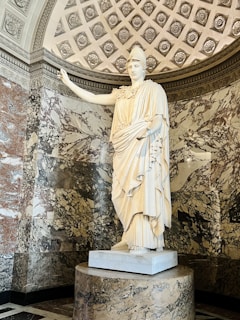 A classical marble statue stands in an ornate alcove. The statue features a draped figure, possibly a goddess or regal figure, with an arm outstretched. The alcove is adorned with intricate patterns and marble inlays in varied tones of gray, white, and brown.