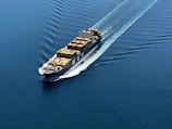 A cargo ship is sailing through a vast expanse of blue water, carrying numerous colorful shipping containers. The ship leaves a trail of waves behind, slicing smoothly through the calm sea.