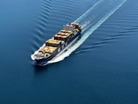 A cargo ship is sailing through a vast expanse of blue water, carrying numerous colorful shipping containers. The ship leaves a trail of waves behind, slicing smoothly through the calm sea.