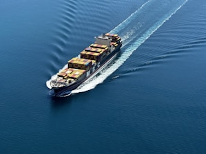 A cargo ship is sailing through a vast expanse of blue water, carrying numerous colorful shipping containers. The ship leaves a trail of waves behind, slicing smoothly through the calm sea.