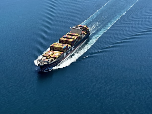 A cargo ship is sailing through a vast expanse of blue water, carrying numerous colorful shipping containers. The ship leaves a trail of waves behind, slicing smoothly through the calm sea.