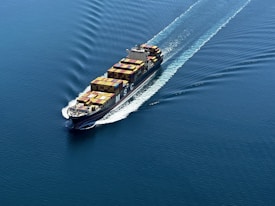 A cargo ship is sailing through a vast expanse of blue water, carrying numerous colorful shipping containers. The ship leaves a trail of waves behind, slicing smoothly through the calm sea.