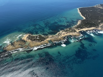 A large, lush island with a rugged coastline extends into the ocean, surrounded by clear turquoise waters. The landscape features diverse terrains with patches of greenery and sandy beaches. Waves crash against the shoreline, creating white foam.