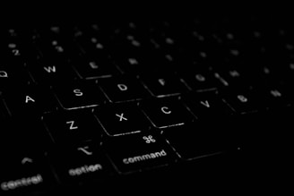 A close-up view of a black computer keyboard with white lettering. The focus is on the keys labeled A, S, D, X, and C, along with special command keys. The lighting is low, creating a high-contrast effect and emphasizing the texture of the keys.
