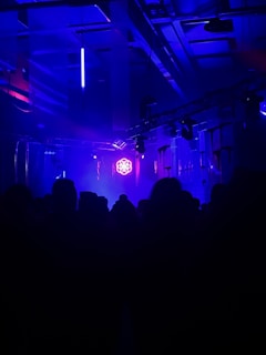 A dark room filled with silhouetted figures, illuminated by vivid blue and purple lighting. The ceiling features exposed structures and hanging fixtures. Bright neon lights cast an atmospheric glow, creating a dynamic and immersive setting.