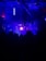 A dark room filled with silhouetted figures, illuminated by vivid blue and purple lighting. The ceiling features exposed structures and hanging fixtures. Bright neon lights cast an atmospheric glow, creating a dynamic and immersive setting.