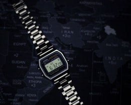 A classic Casio digital wristwatch with a metallic strap is placed on a dark world map. The display shows the time and date, featuring an alarm and water resistance. The map has a dark, subdued color palette, highlighting countries in the Middle East and parts of Asia.