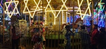 A lively amusement park scene featuring a brightly illuminated carousel with ornate decorations. Several people stand around the carousel, enjoying the festive atmosphere. Stalls nearby display a range of colorful toys and accessories. The background is filled with vivid neon lights, adding to the carnival ambiance.