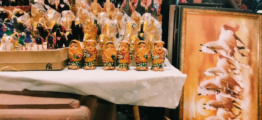 A collection of colorful handcrafted figurines is displayed on a table, with four intricately detailed golden figures in the foreground. Behind them, more figurines wrapped in plastic are visible. To the right, there's a framed artwork showing dynamic horses galloping against a warm background.