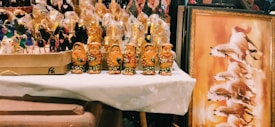A collection of colorful handcrafted figurines is displayed on a table, with four intricately detailed golden figures in the foreground. Behind them, more figurines wrapped in plastic are visible. To the right, there's a framed artwork showing dynamic horses galloping against a warm background.