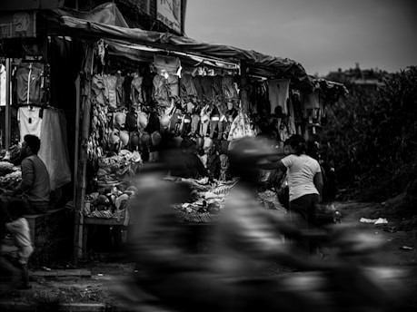 A bustling street market stall displays various items of clothing and accessories, with several people shopping or moving past. The focus is on a man sitting by the stall and a woman browsing through clothes, while the scene captures the motion blur of people passing by swiftly. The setting seems dynamic and lively, with goods hanging overhead and scattered around.