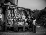 A black and white image captures a bustling outdoor market scene. Several people are gathered around a small shop filled with various clothing items including shirts, bras, and other garments hanging above and displayed on the table. The people appear engaged in conversation, with one woman prominently positioned at the center, looking up.