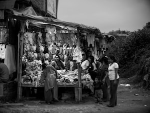 A black and white image captures a bustling outdoor market scene. Several people are gathered around a small shop filled with various clothing items including shirts, bras, and other garments hanging above and displayed on the table. The people appear engaged in conversation, with one woman prominently positioned at the center, looking up.