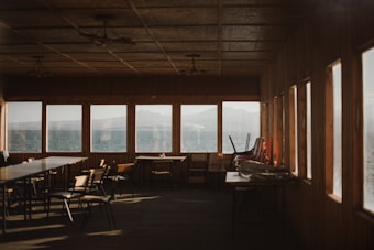 The image depicts an empty wooden room with a row of large windows overlooking a calm body of water and distant mountains. The room contains tables and chairs, some of which are stacked. The lighting is warm and soft, suggesting late afternoon or early evening.