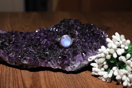 A cluster of dark amethyst crystals sits on a wooden surface, exhibiting a rich purple hue. Resting on the crystals is a silver ring with a luminous blue stone, adding an elegant touch. In the foreground, a collection of delicate white flowers with green stems provides a contrasting visual element.