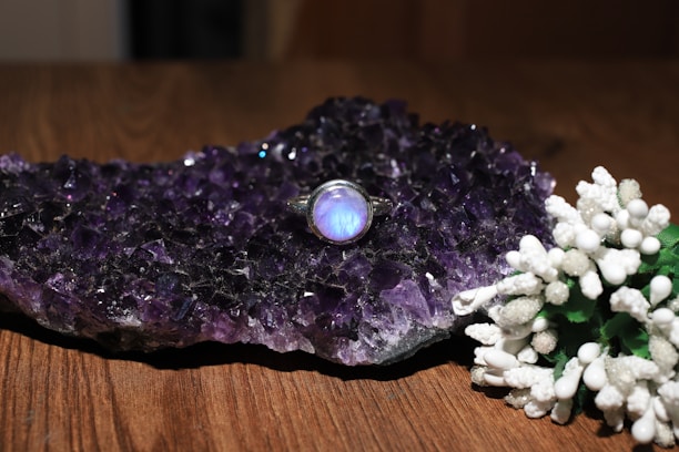 A cluster of dark amethyst crystals sits on a wooden surface, exhibiting a rich purple hue. Resting on the crystals is a silver ring with a luminous blue stone, adding an elegant touch. In the foreground, a collection of delicate white flowers with green stems provides a contrasting visual element.