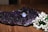 A cluster of dark amethyst crystals sits on a wooden surface, exhibiting a rich purple hue. Resting on the crystals is a silver ring with a luminous blue stone, adding an elegant touch. In the foreground, a collection of delicate white flowers with green stems provides a contrasting visual element.