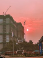 A construction site features prominently with a partially built high-rise covered in green construction netting. Cranes are visible above the building. The sky is illuminated by the setting or rising sun, casting an orange-pink hue over the scene. Several vehicles, including a truck and cars, are present on the street in front of the site. Trees and a fuel station sign are also noticeable in the foreground.