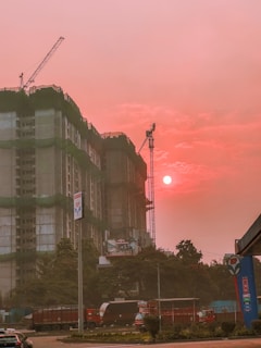 A construction site features prominently with a partially built high-rise covered in green construction netting. Cranes are visible above the building. The sky is illuminated by the setting or rising sun, casting an orange-pink hue over the scene. Several vehicles, including a truck and cars, are present on the street in front of the site. Trees and a fuel station sign are also noticeable in the foreground.