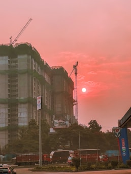 A construction site features prominently with a partially built high-rise covered in green construction netting. Cranes are visible above the building. The sky is illuminated by the setting or rising sun, casting an orange-pink hue over the scene. Several vehicles, including a truck and cars, are present on the street in front of the site. Trees and a fuel station sign are also noticeable in the foreground.