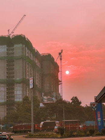 A construction site features prominently with a partially built high-rise covered in green construction netting. Cranes are visible above the building. The sky is illuminated by the setting or rising sun, casting an orange-pink hue over the scene. Several vehicles, including a truck and cars, are present on the street in front of the site. Trees and a fuel station sign are also noticeable in the foreground.