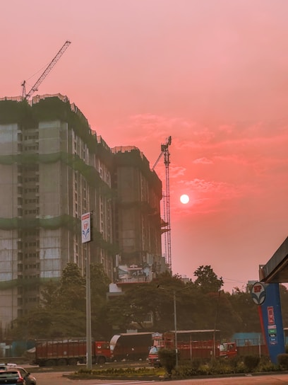 A construction site features prominently with a partially built high-rise covered in green construction netting. Cranes are visible above the building. The sky is illuminated by the setting or rising sun, casting an orange-pink hue over the scene. Several vehicles, including a truck and cars, are present on the street in front of the site. Trees and a fuel station sign are also noticeable in the foreground.