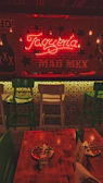 A warmly lit interior of a restaurant featuring a neon sign that reads 'Taquería' and 'MAD MEX'. The decor includes hanging light bulbs, green and white bar stools, a wooden table set with painted plates, and Mexican-themed graphics on the walls. The ambiance suggests a cozy and inviting atmosphere.