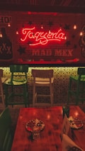 A warmly lit interior of a restaurant featuring a neon sign that reads 'Taquería' and 'MAD MEX'. The decor includes hanging light bulbs, green and white bar stools, a wooden table set with painted plates, and Mexican-themed graphics on the walls. The ambiance suggests a cozy and inviting atmosphere.