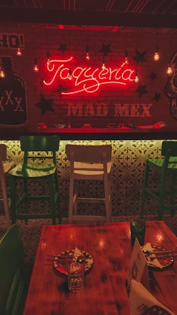 A warmly lit interior of a restaurant featuring a neon sign that reads 'Taquer&iacute;a' and 'MAD MEX'. The decor includes hanging light bulbs, green and white bar stools, a wooden table set with painted plates, and Mexican-themed graphics on the walls. The ambiance suggests a cozy and inviting atmosphere.