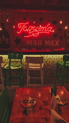 A warmly lit interior of a restaurant featuring a neon sign that reads 'Taquería' and 'MAD MEX'. The decor includes hanging light bulbs, green and white bar stools, a wooden table set with painted plates, and Mexican-themed graphics on the walls. The ambiance suggests a cozy and inviting atmosphere.