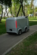 A small, futuristic-looking autonomous vehicle is traveling on a paved path through a lush park. The vehicle has a sleek gray exterior with red vertical lights on the rear. Tall trees with green foliage surround the path, and the ground is covered with grass and sparse vegetation.